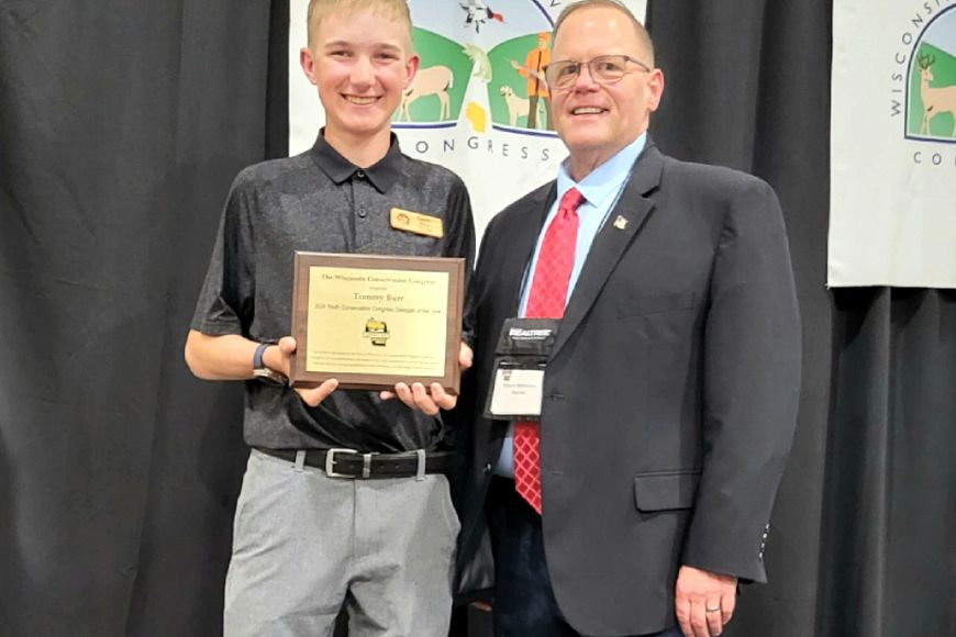 Two individuals standing in front of a banner that reads "Wisconsin Conservation Congress", holding an award plaque together and smiling at the camera.