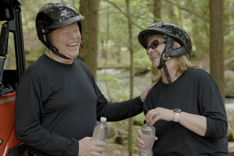 Two UTV riders take a break outside their vehicle to talk and drink water.