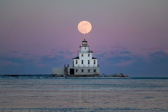 The huge full moon glows brightly and is perfectly centered above the spire of the Manitowoc Lighthouse. The sky transitions from a pale pink to dark blue at the horizon line, where it merges with the icy Lake Michigan waters. A light skim of chunky ice covering the water reflects the colors of the sky above.