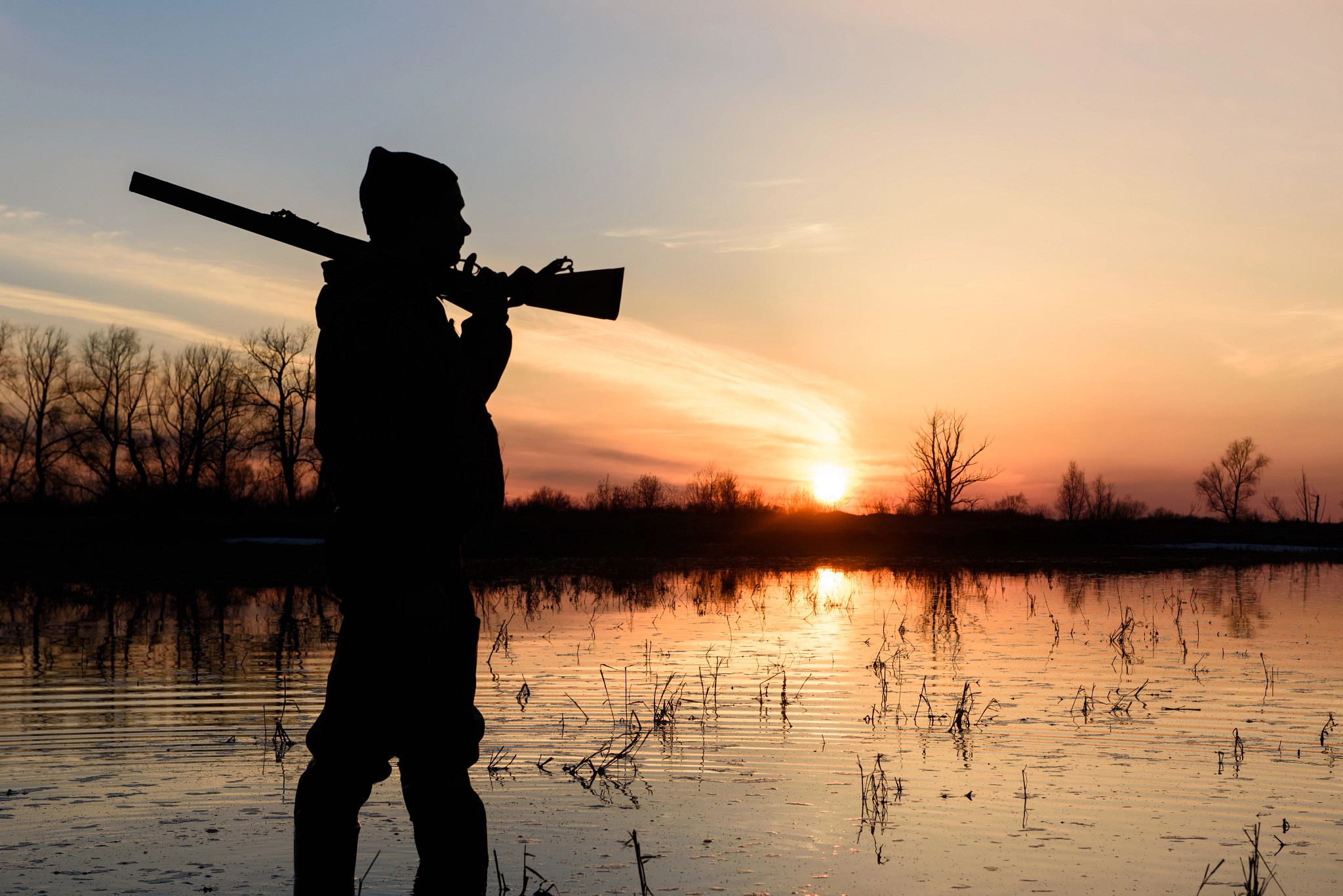 A waterfowl hunter with a gun over their shoulder silhouetted at sunset. 