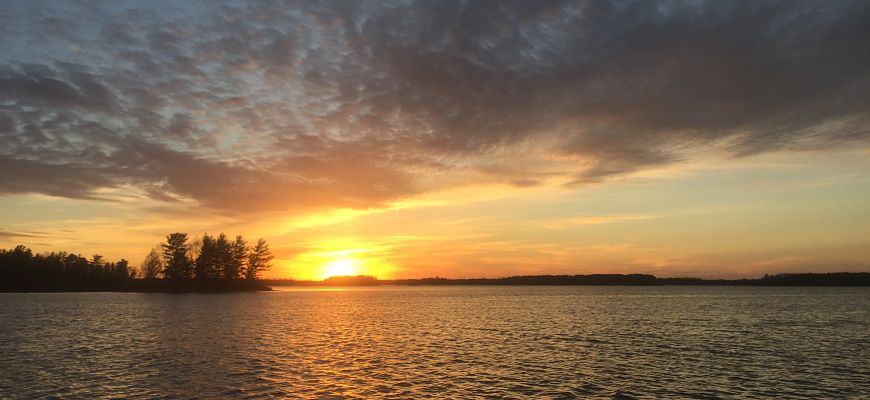Sun setting below the horizon under a grey and blue sky with calm waters in the foreground.