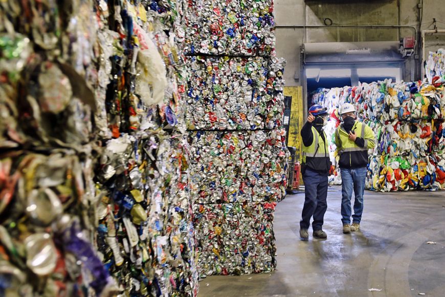 Two recycling plant workers walk through a warehouse lined with bales of recycled plastic.