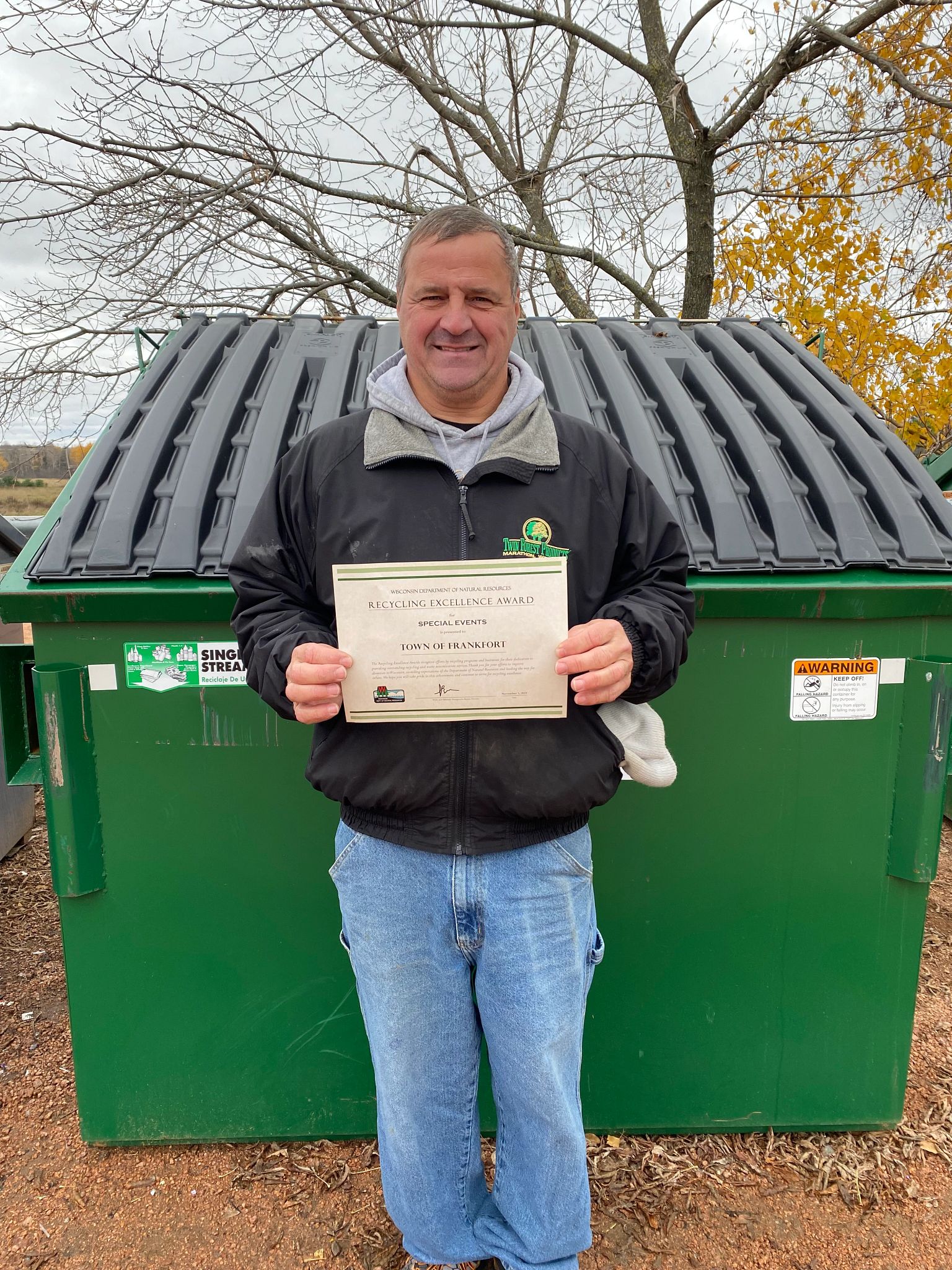 A Town of Frankfort team member stands in front of a green recycling dumpster while holding the town's 2023 Wisconsin Recycling Excellence Award certificate from the DNR.