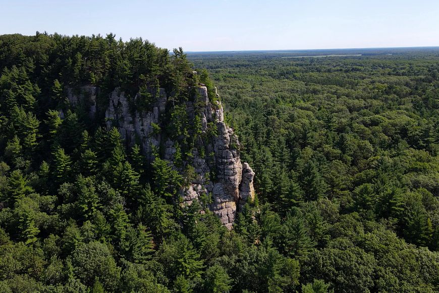 A drone photo of the bluff at Roche-A-Cri State Park on a sunny day. The bluff is covered in pine trees with blue sky above.