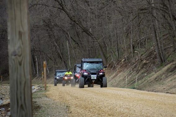Three UTVs and one ATV ride in a line along a trail bordered by tall trees.