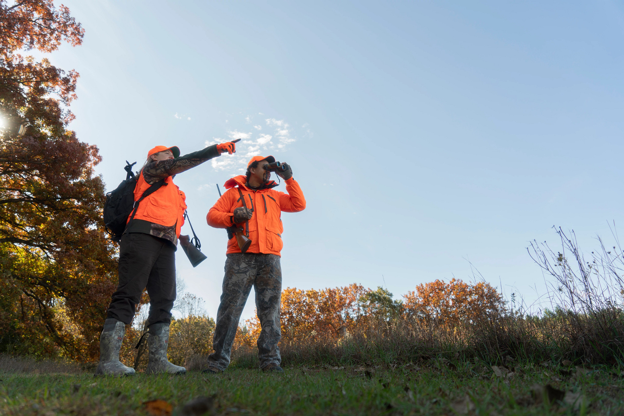 A couple wearing blaze orange scouts a field with binoculars. 