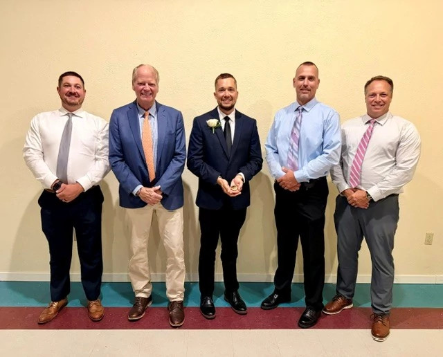 five individuals stand side-by-side in formal attire in front of a wall