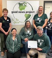Six Good News Project representatives pose with their award in front of a Good News Project sign.