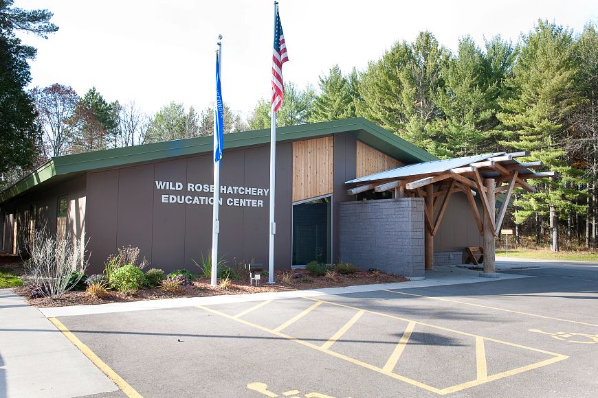 A gray building with a green roof has the words "Wild Rose Hatchery Education Center" on its siding. In front of the building, the American and Wisconsin State flags are seen flying.