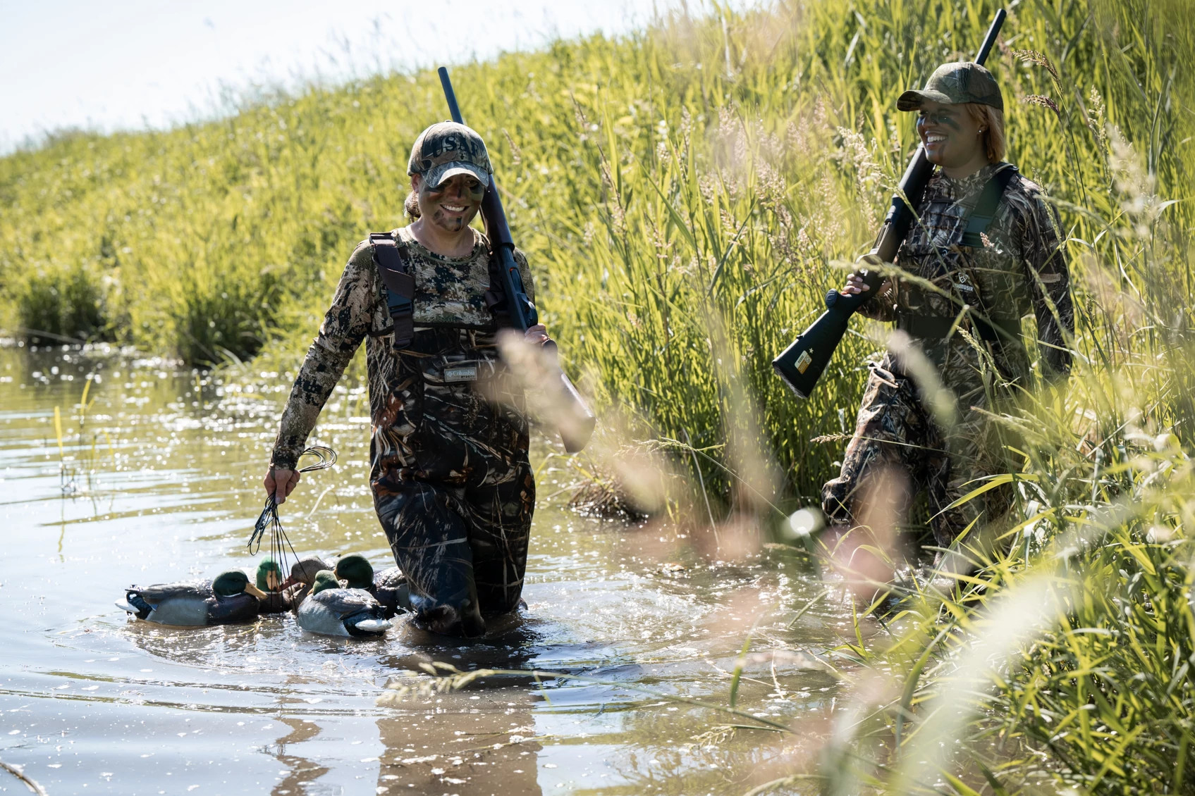 Two individuals in camouflage hunting gear wading through a marshy area carrying decoys.