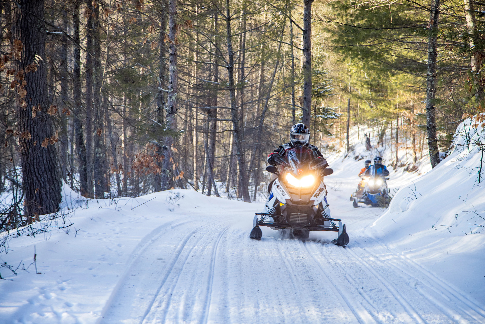 three people riding on snowmobiles with helmets on a snowy trail through a forest