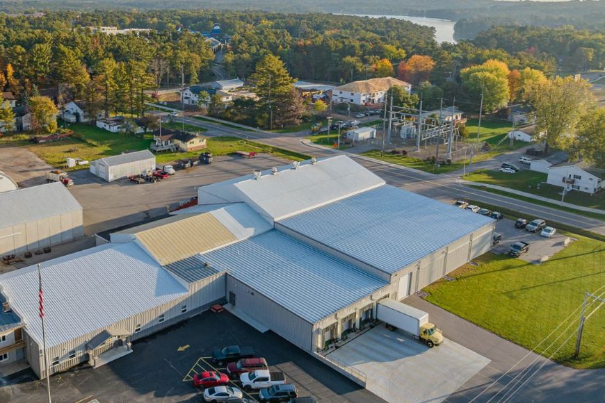 A drone photo of a large building/warehouse with trucks and cars on the outside and forest in the back