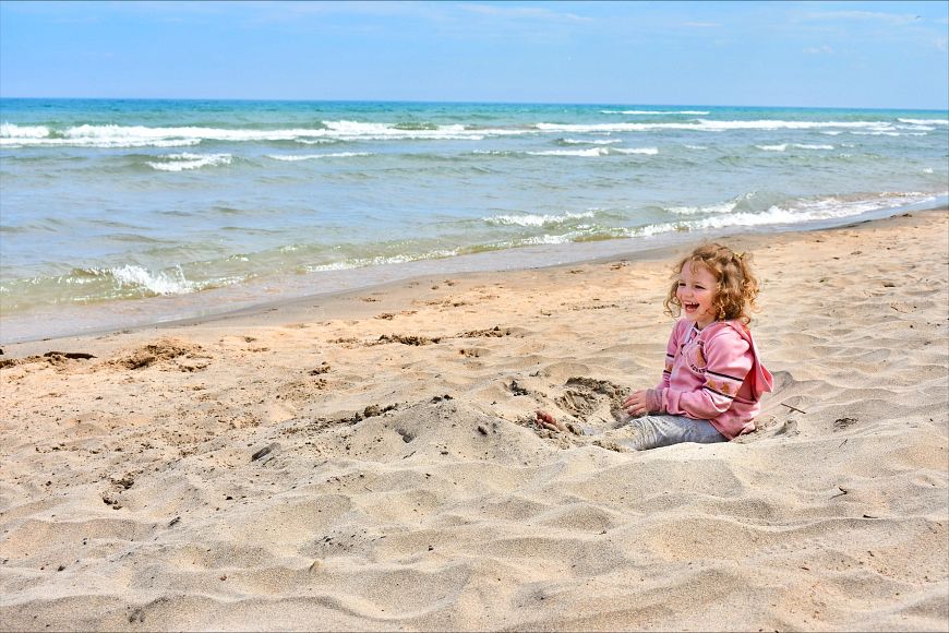 A child is sitting on a sandy beach, smiling joyfully with the ocean waves in the background on a bright sunny day.