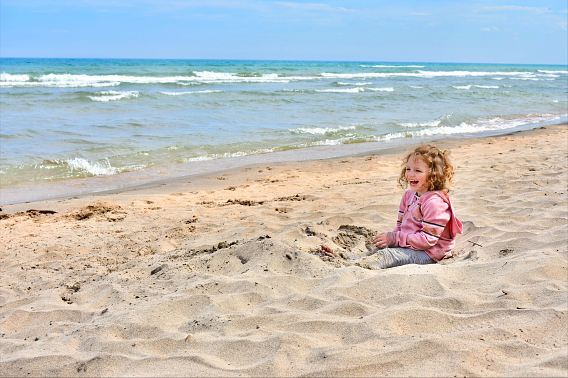 A young girl laughs while sitting on a sandy beach with her feet partially buried in the sand. Small waves break near the beach.