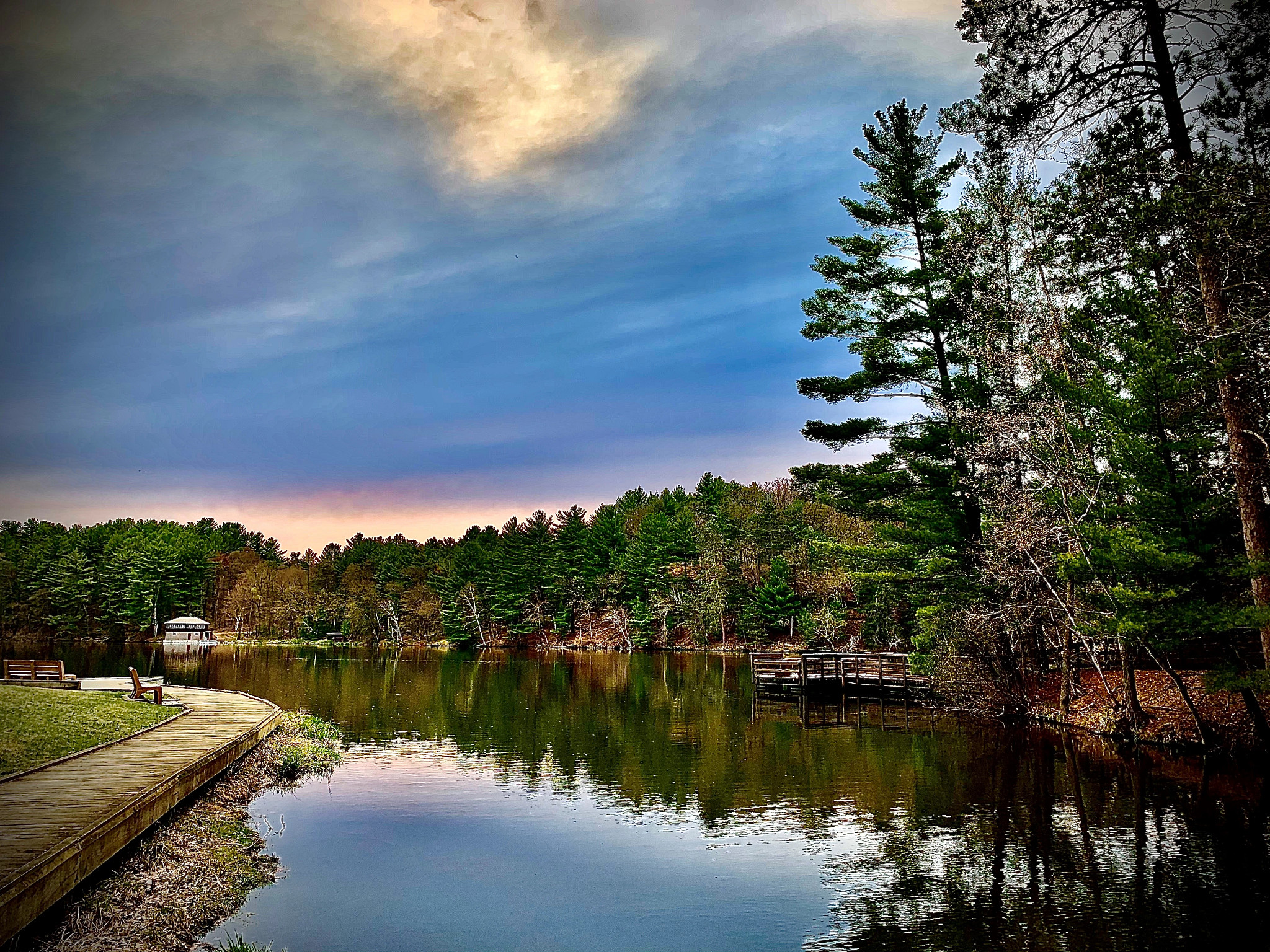 A view of the Mirror Lake board walk on the left hand side. The lake is shown along the board walk and the tree line following the lake with a cloudy blue sky.