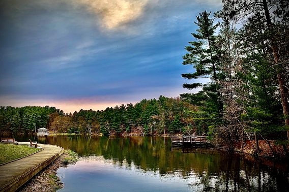 A view of the Mirror Lake board walk on the left hand side. The lake is shown along the board walk and the tree line following the lake with a cloudy blue sky.