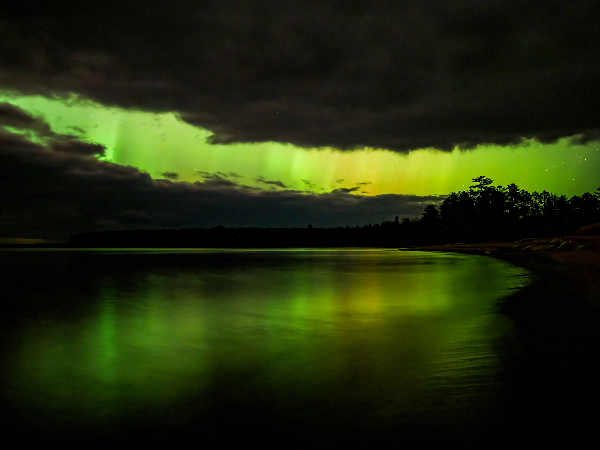 Shades of green, yellow and purple from the Aurora Borealis illuminate the night sky and cast their reflections on still waters of the crescent-shaped bay. Moody dark clouds take up much of the sky, except for a large gap between them, where the aurora beams through along with a scattering of stars. Pine and spruce trees are silhouetted against the aurora’s colors.