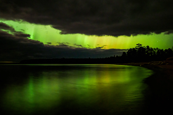 Shades of green, yellow and purple from the Aurora Borealis illuminate the night sky and cast their reflections on still waters of the crescent-shaped bay. Moody dark clouds take up much of the sky, except for a large gap between them, where the aurora beams through along with a scattering of stars. Pine and spruce trees are silhouetted against the aurora’s colors.