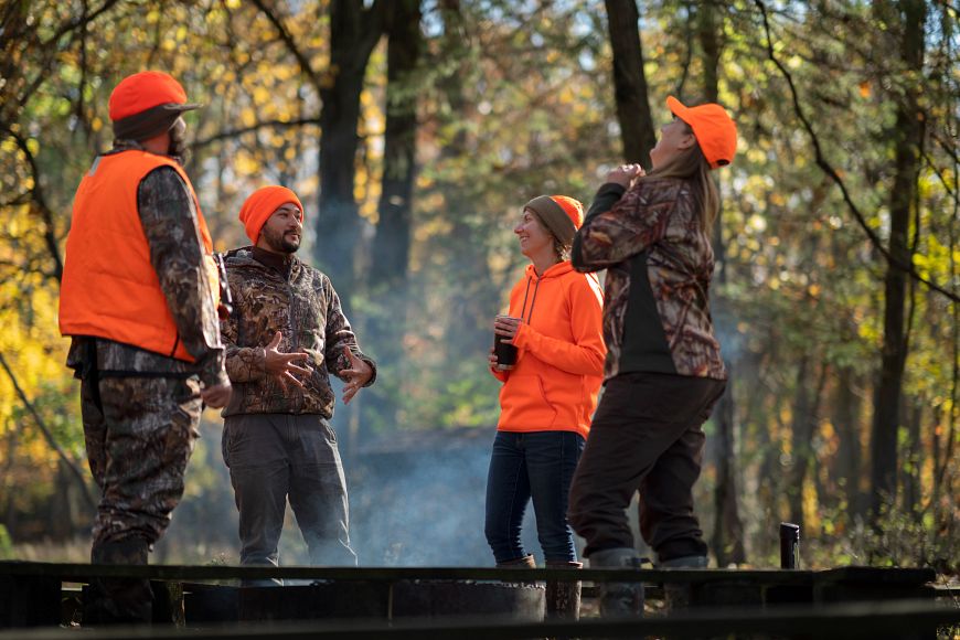 A group of four hunters, hanging out and laughing in the woods around a campfire.