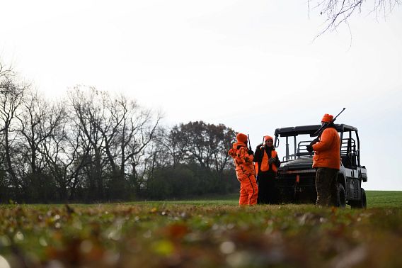 Three hunters, all in blaze orange, stand around their UTV at the edge of a wooded area.