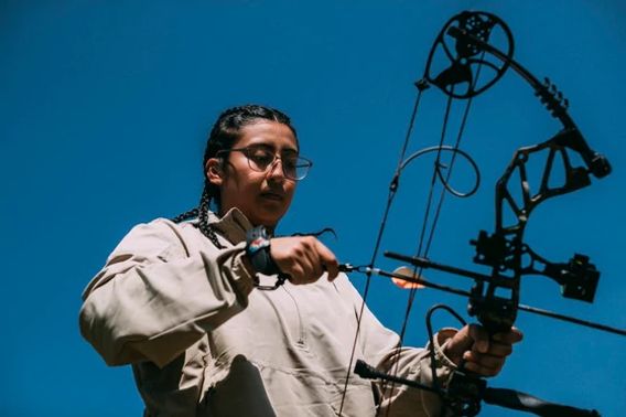 A young person wearing a tan jacket holds examines a bow before drawing and taking aim. 