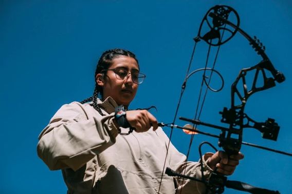 A young person wearing a tan jacket holds examines a bow before drawing and taking aim.