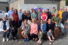 A large group of volunteers pose outside for a photo, with people in the front row sitting in donated chairs and a sofa.