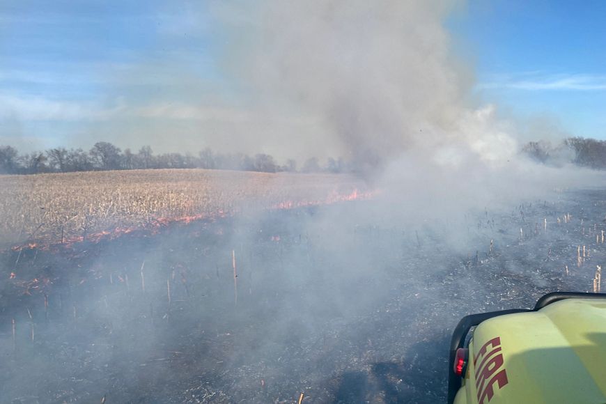 The front of a fire truck is seen in front of a burning field giving off thick gray smoke.