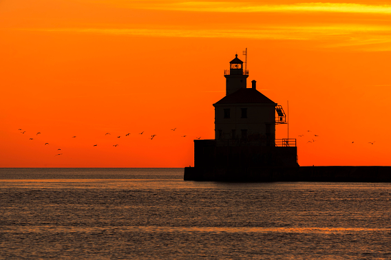The Wisconsin Point Lighthouse is in silhouette against a brilliant orange sunrise. A flock of seagulls stretches along the horizon above the calm waters.