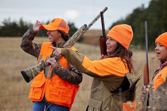 Three women, all wearing Blaze Orange clothing, hunt together in a cornfield during the fall. One woman points to a target outside of the frame. 