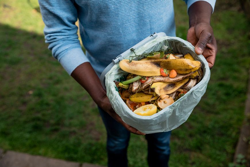 A person holding a small trash can full of compostable food scraps like banana peels, egg shells and lemon rinds.