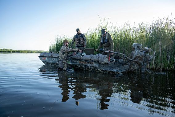 Three waterfowl hunters and their hunting dog on a boat alongside tall cattails.