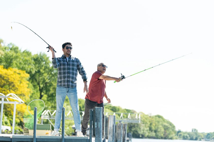 Two men stand on a metal dock on a bright, sunny day, casting fishing rods. 