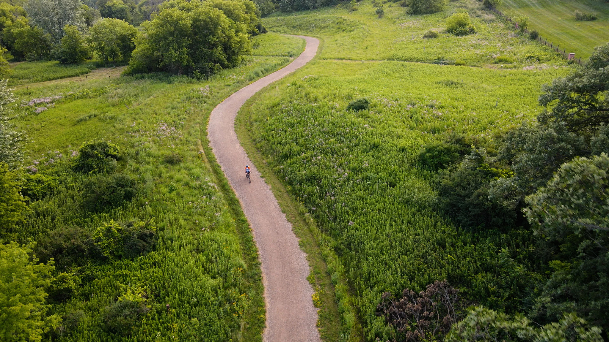 Aerial photo of the trail with a cyclist on it winding through green fields with some spotty trees.