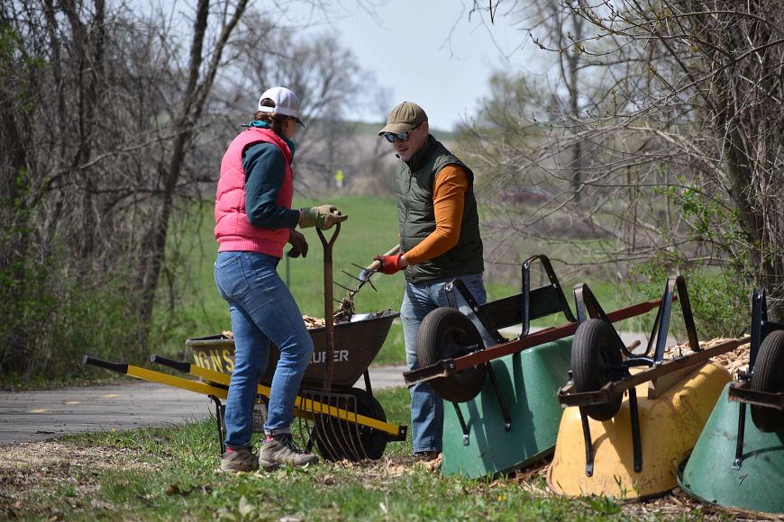 two people shoveling dirt into a wheelbarrow in a grassy area near woods