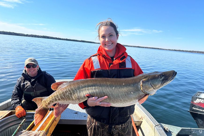 a woman holding a musky on a boat on a lake with a man in the background
