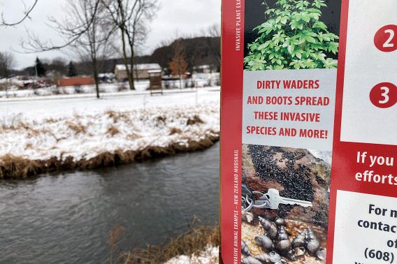 A closeup of a sign saying, "Dirty waders and boots spread these invasive species and more!" in front of a stream with snow on the ground.