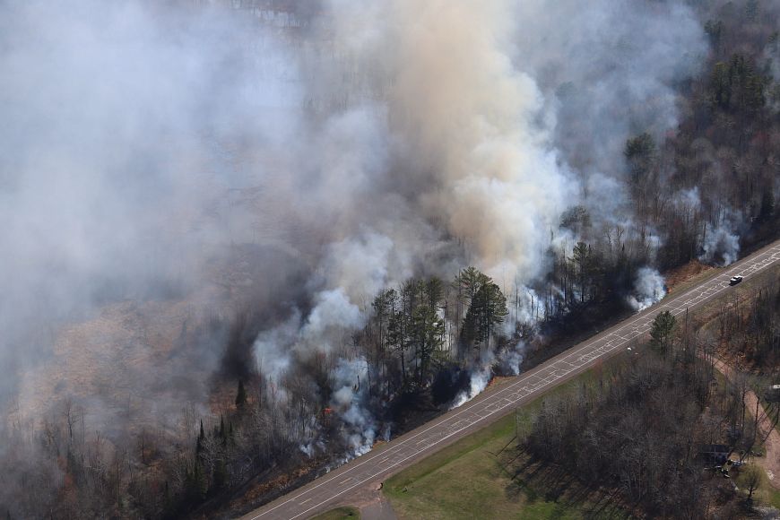 An aerial view of a wildfire burning in the ditch alongside a country road. White and gray smoke fills the air.