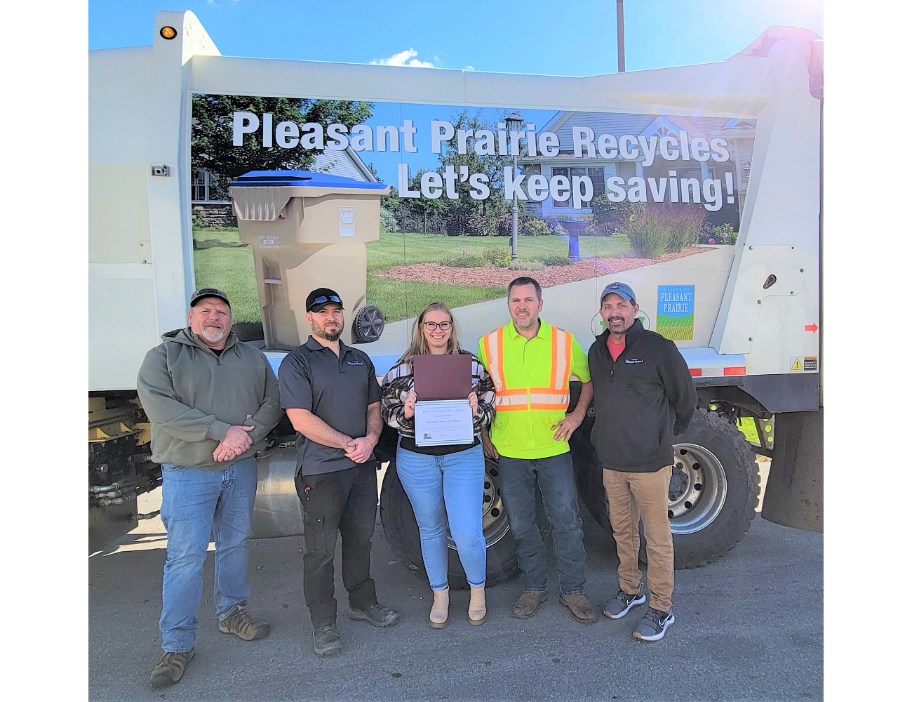 Five Pleasant Prairie village representatives pose with their award outside next to a recycling truck. The truck has a printed sign on the side with a picture of a recycling can on the side of the road and the phrase 'Pleasant Prairie recycles. Let's keep saving!'