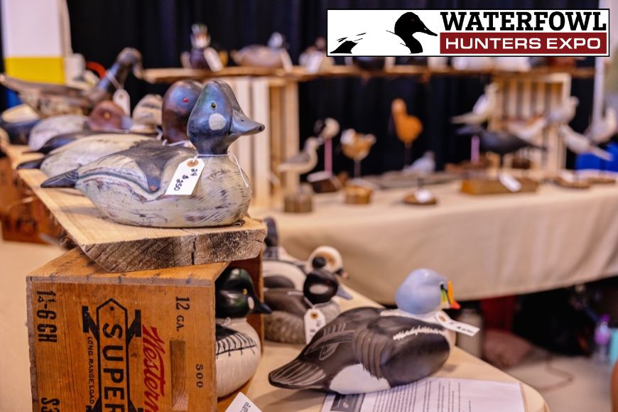 Wooden decoy ducks for sale on a table with a logo in the upper right corner with a black-and-white duck and the words "Waterfowl Hunters Expo."