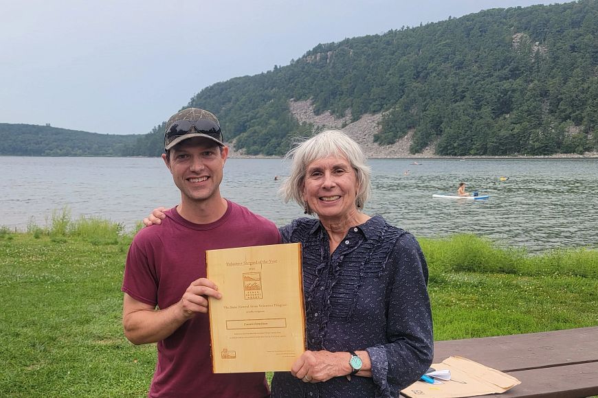 two people holding up a wooden plaque near a picnic table in front of Devils Lake
