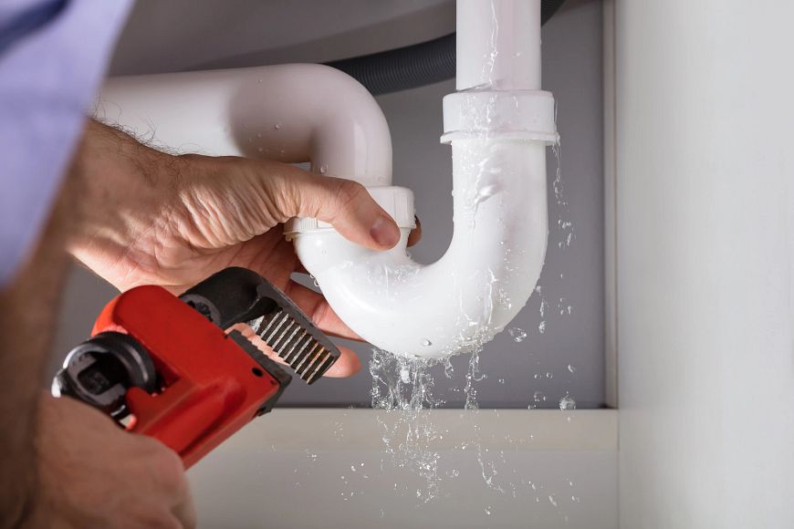 Someone uses a red pipe wrench to fix a leaky PVC pipe with water gushing out. 