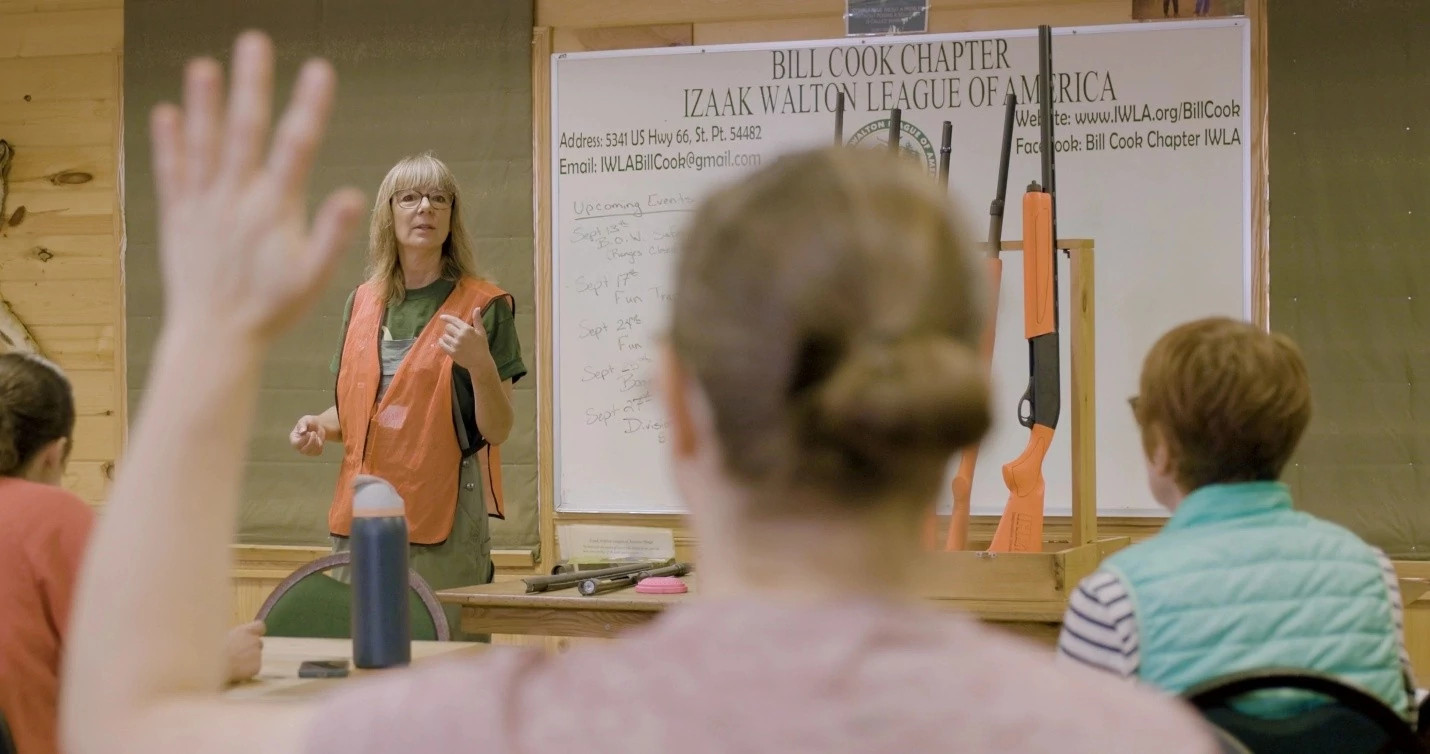 A person in an orange safety vest stands at the front of a room, gesturing towards a board with the name "Bill Cook Chapter Izaak Walton League of America." Several people are seated, one with a hand raised. A pair of rifles is mounted on the board.