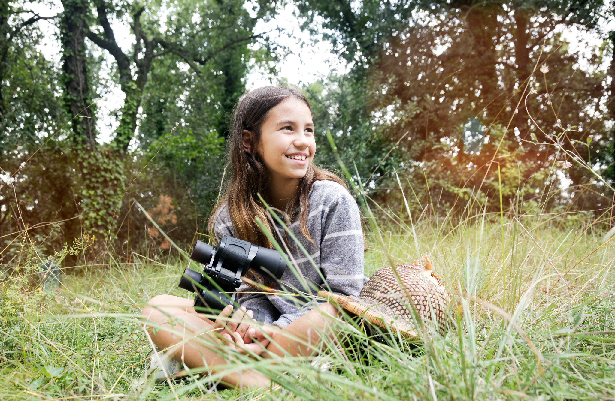 A young girl with long brown hair sits in the grass holding binoculars and smiling into the distance. 