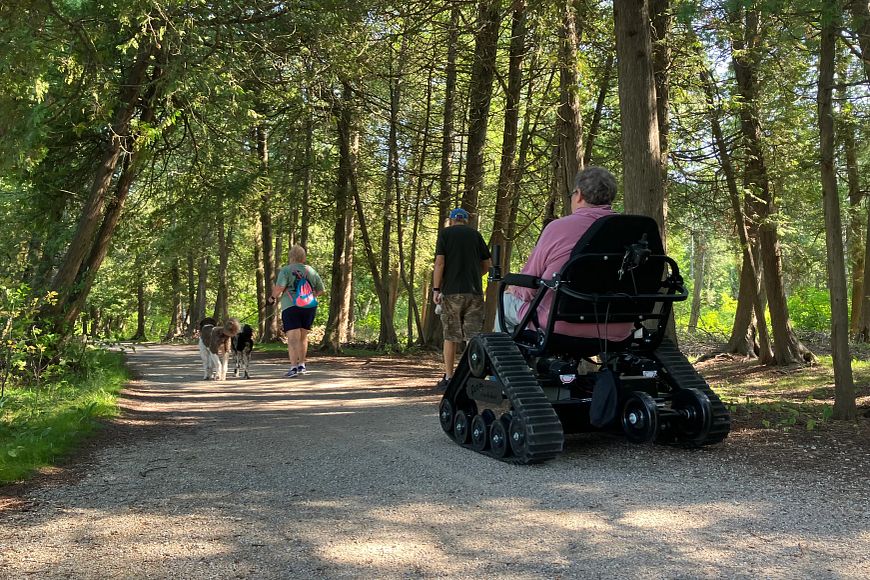 An individual using an all-terrain wheelchair follows two people walking with a dog on a tree-lined path.